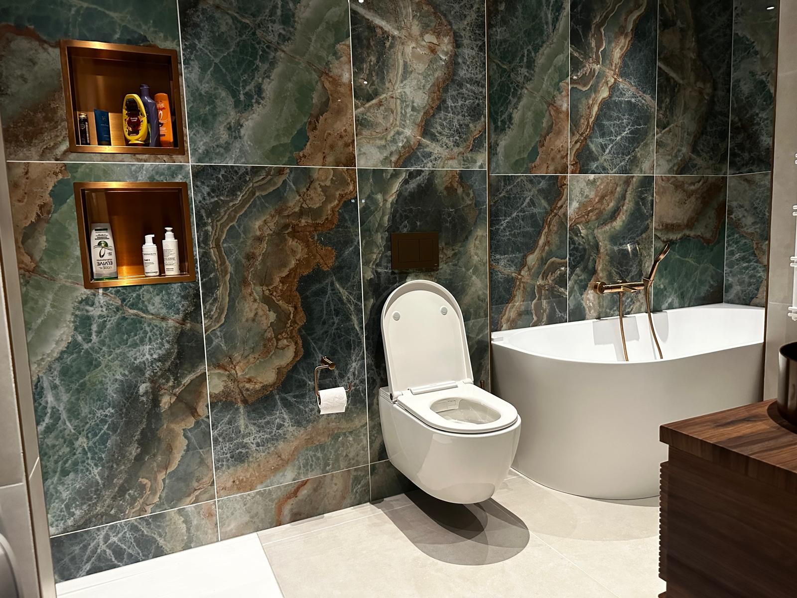 Badkamer in Duivendrecht met groen-bruine natuursteenlook tegelwand, verlichte nissen, hangend toilet en vrijstaand bad met bronskleurige kraan
