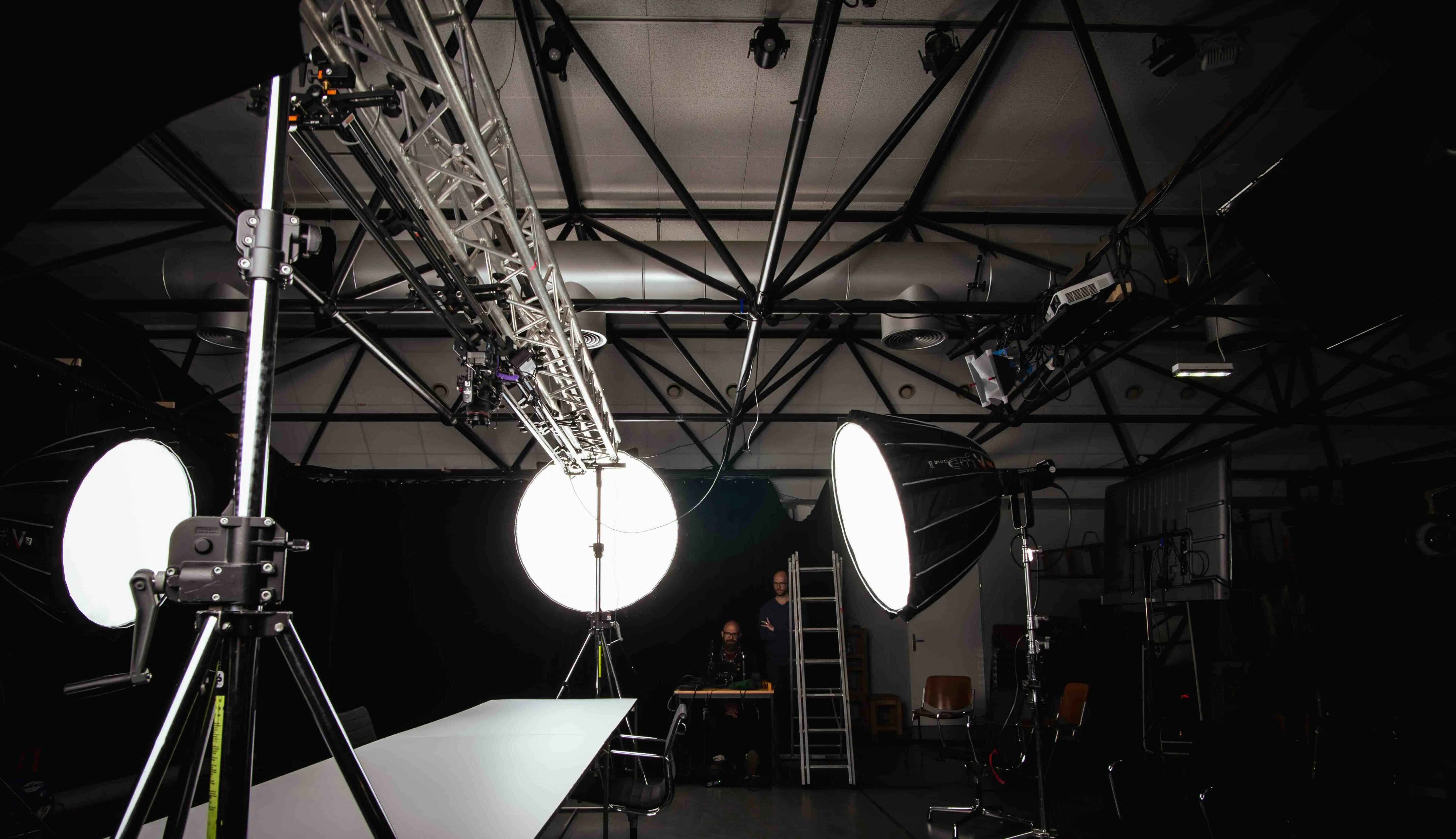 Fotostudio mit drei großen, leuchtenden Softbox-Leuchten, einem langen weißen Tisch, zwei Personen und einer Leiter im Hintergrund.