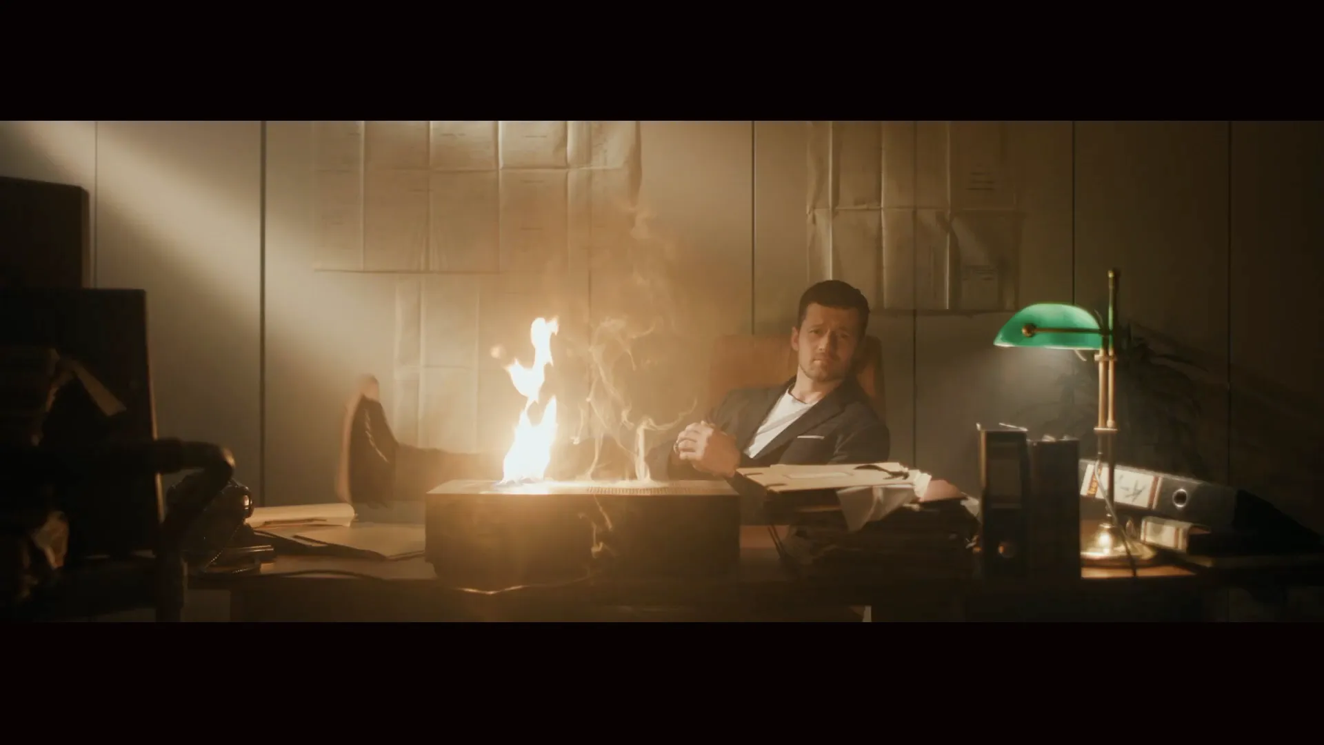 Mann in Anzug sitzt in Büro mit Füßen auf Schreibtisch, auf dem ein Feuer lodert.