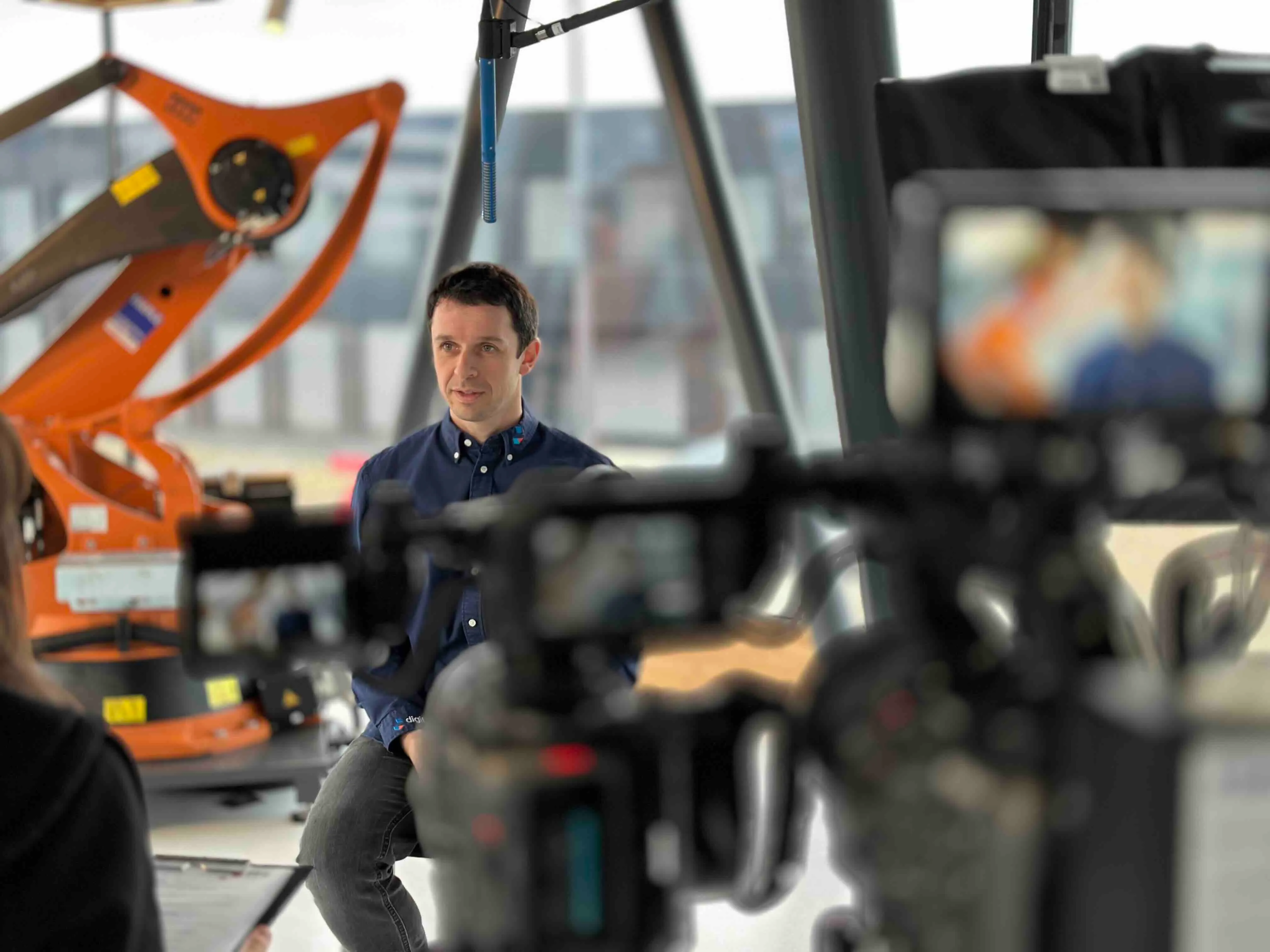 Ein Mann in einem dunkelblauen Hemd wird in einem Industrieumfeld vor einer Kamera interviewt, im Hintergrund ist ein oranger Industrieroboter sichtbar.