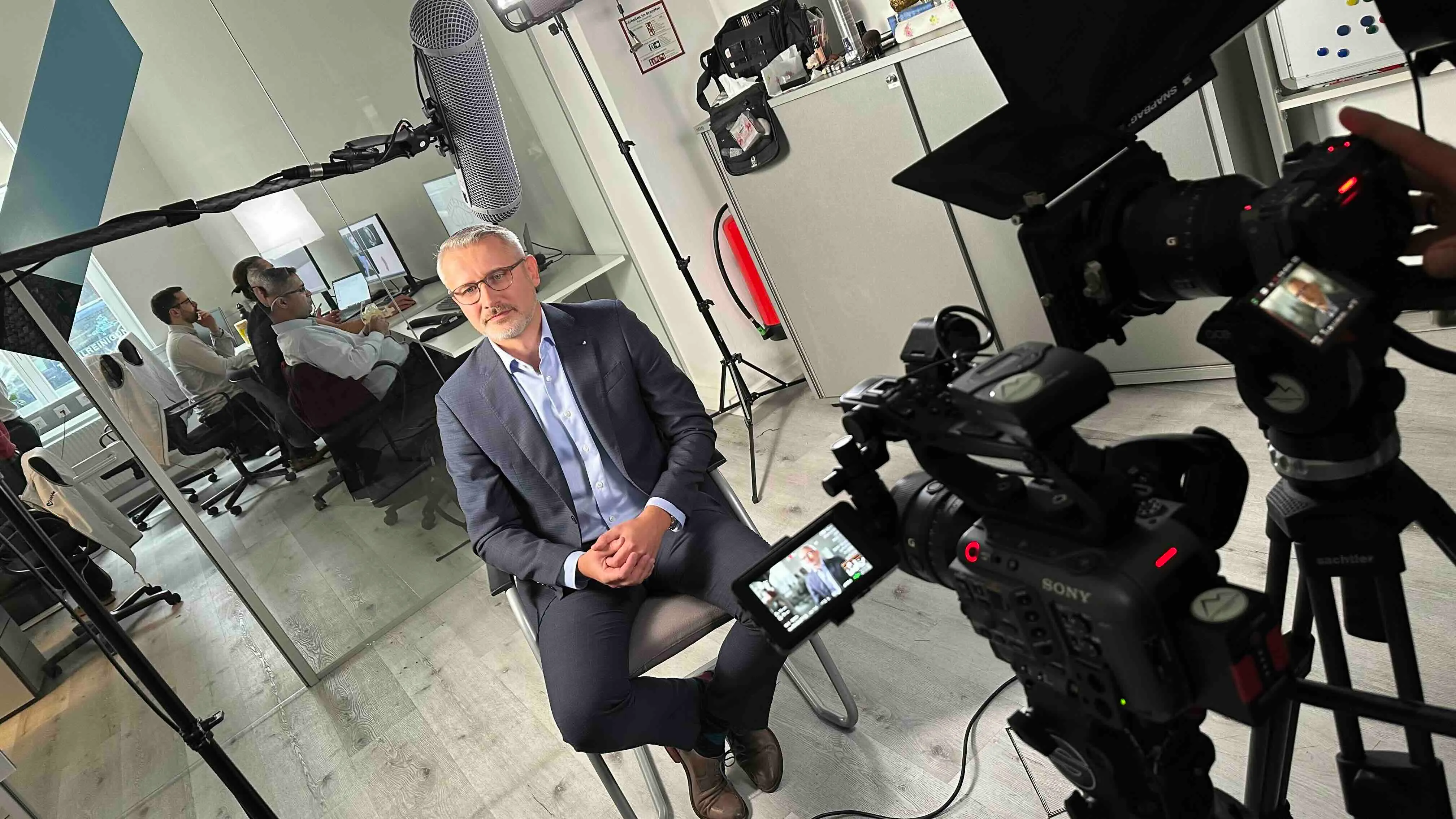 Ein Mann im Anzug sitzt auf einem Stuhl und wird in einem Büro von einer Kamera und Mikrofon für ein Video aufgenommen.