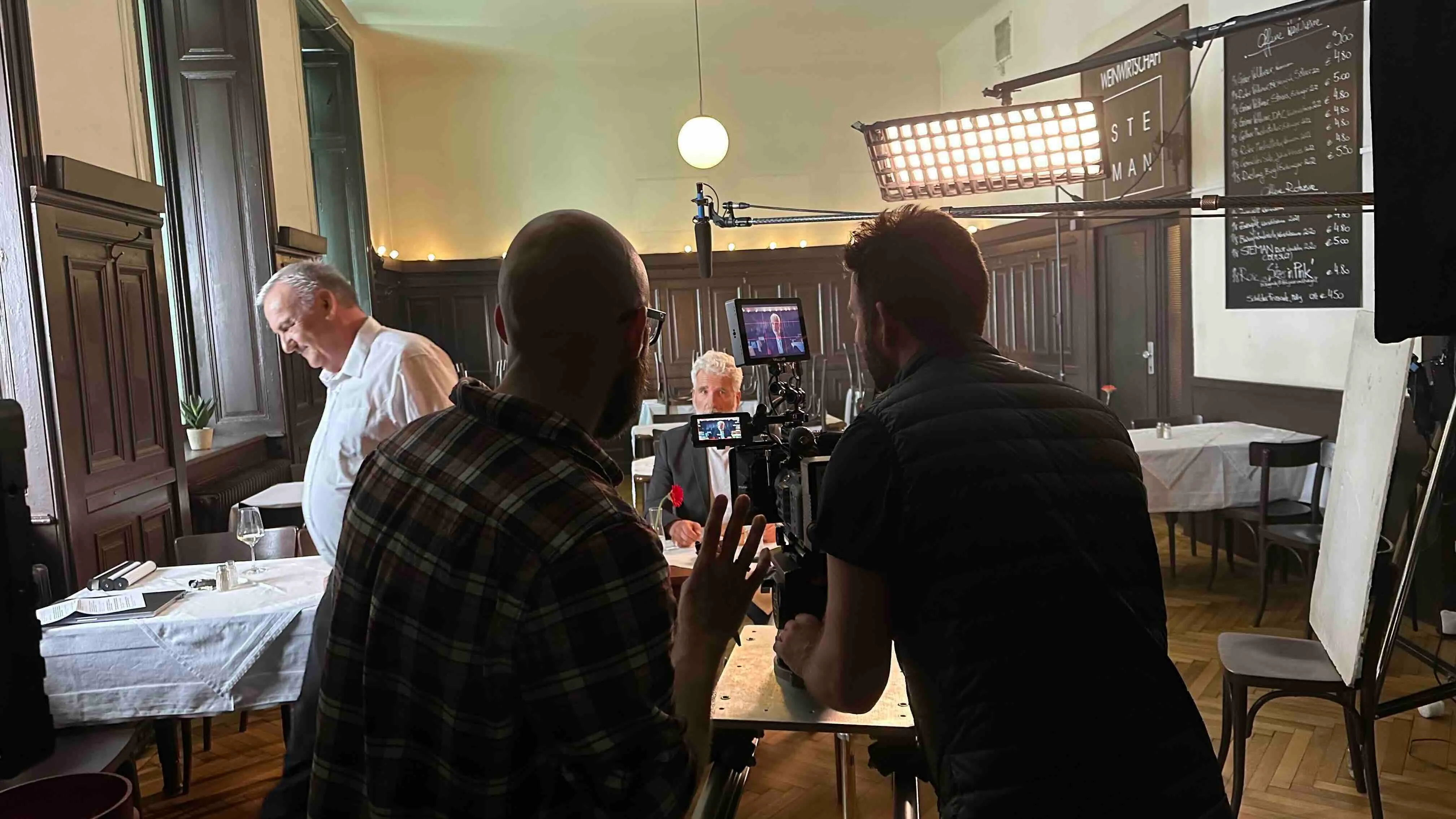 Dreh eines Interviews in einem Café mit einem Mann in Anzug am Tisch, zwei Kameraleuten und professioneller Beleuchtung.