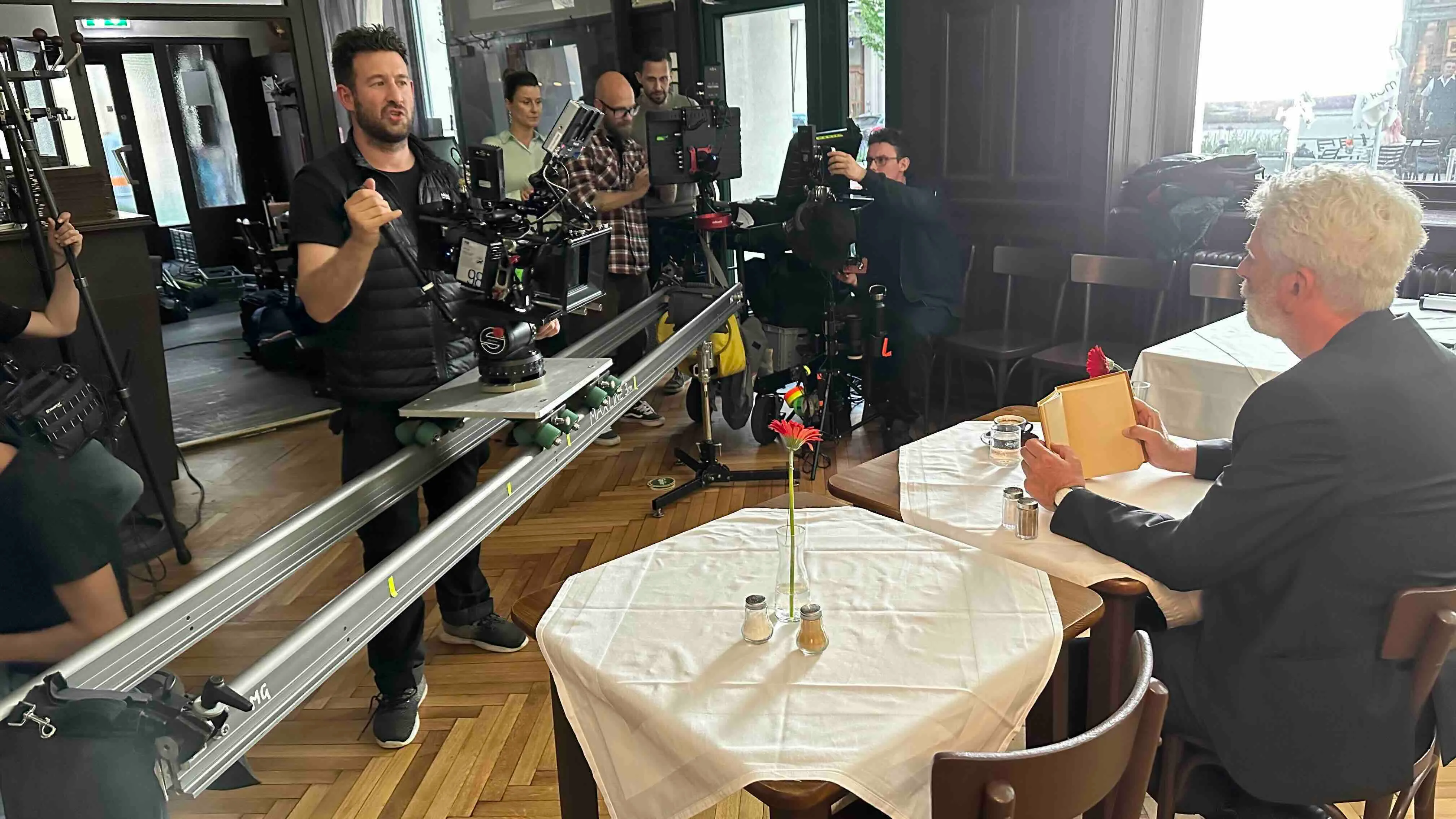 Filmteam richtet Kameras und Technik in einem Restaurant ein, während ein Mann mit grauen Haaren an einem Tisch sitzt und ein Buch hält.