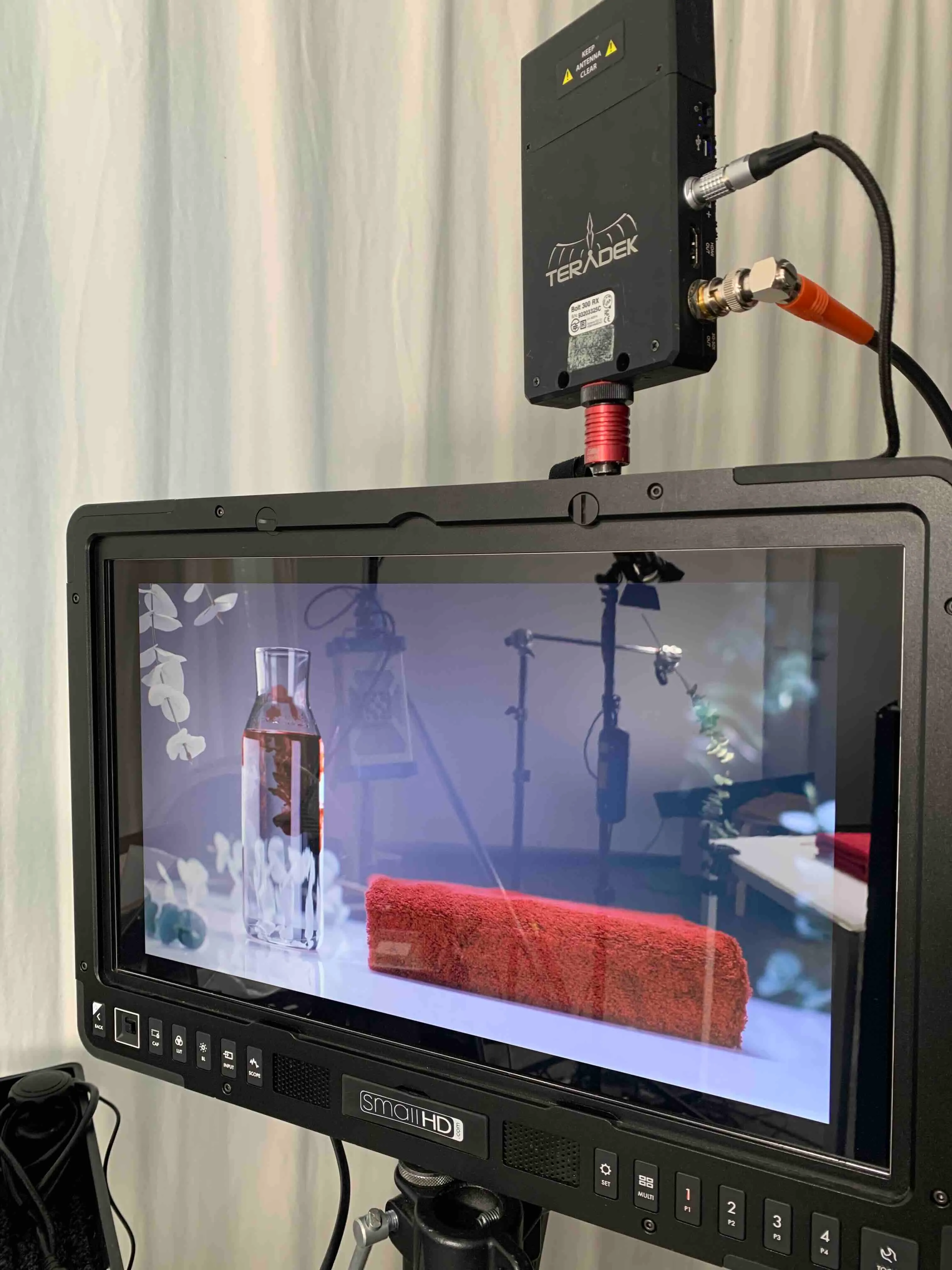 Ein schwarzer SmallHD Monitor zeigt eine Szene mit einer Glasflasche und einem roten Handtuch auf einem weißen Tisch, mit Studiolicht- und Kamerastativ im Hintergrund.