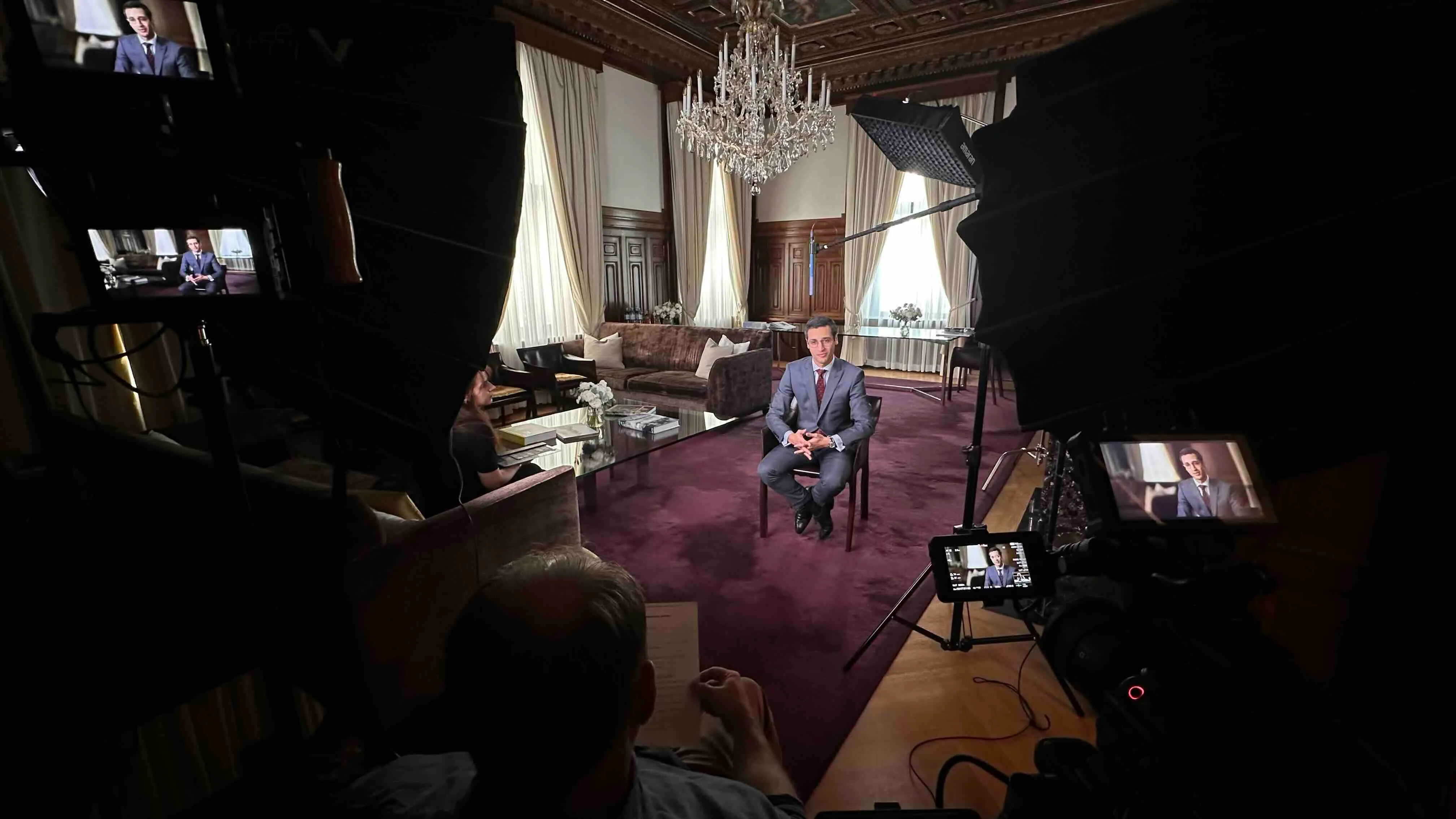 Ein Mann im Anzug sitzt in einem eleganten Raum mit Kronleuchter und Holzvertäfelung, umgeben von Kameras und Beleuchtung für ein Interview.