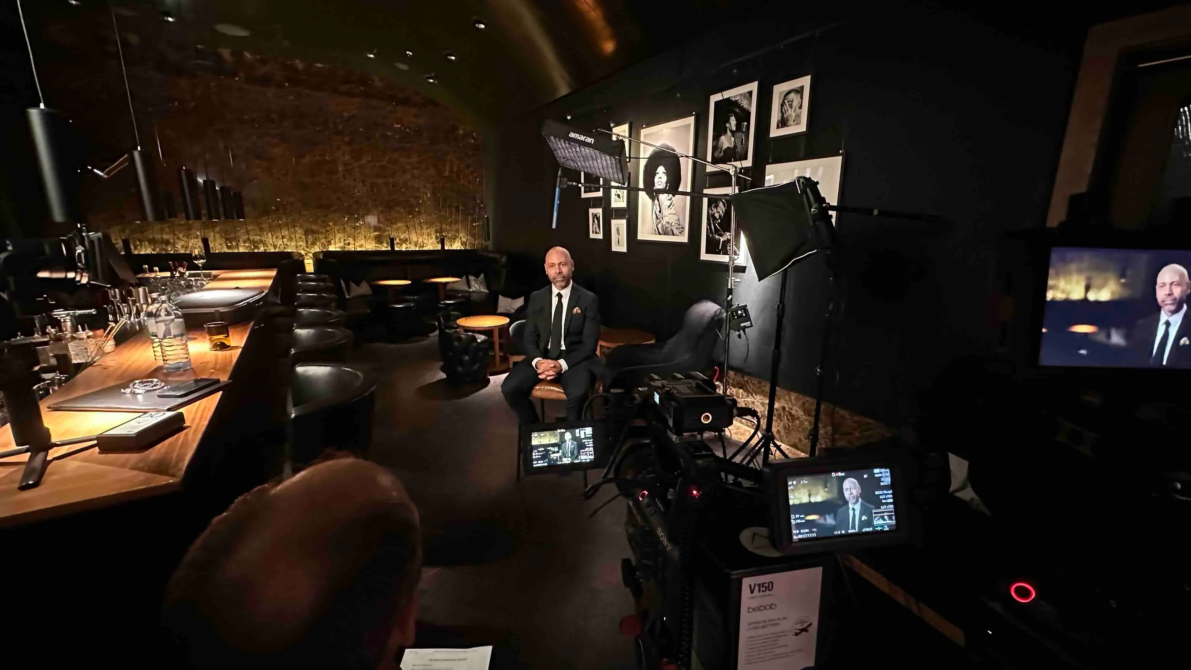 Ein Mann im Anzug sitzt für ein Interview in einem stilvollen, dunklen Raum mit Kameraausrüstung und Beleuchtung.