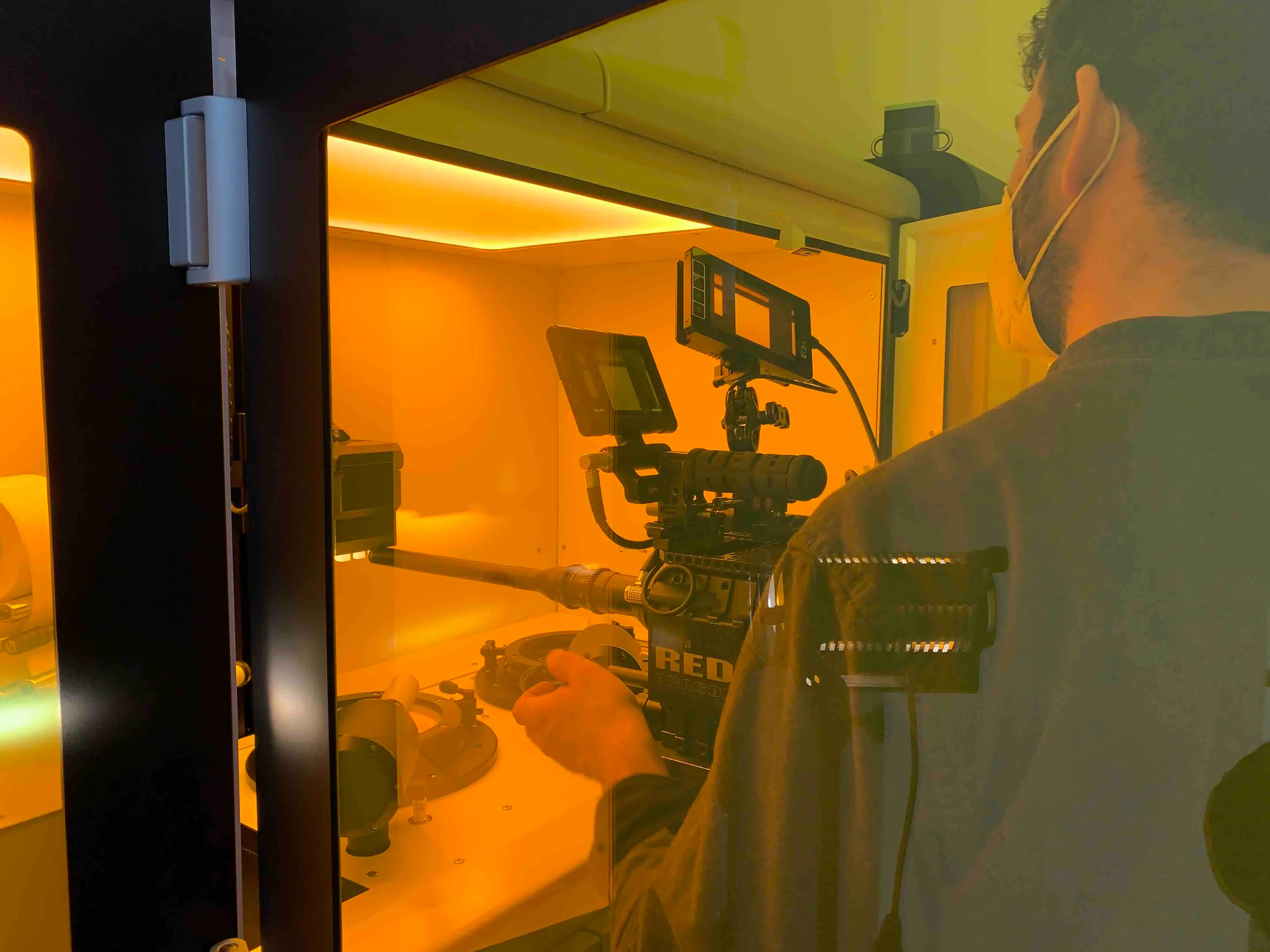 Person mit Mund-Nasen-Schutz bedient professionelle RED-Kamera hinter einer gelb getönten Glasscheibe.