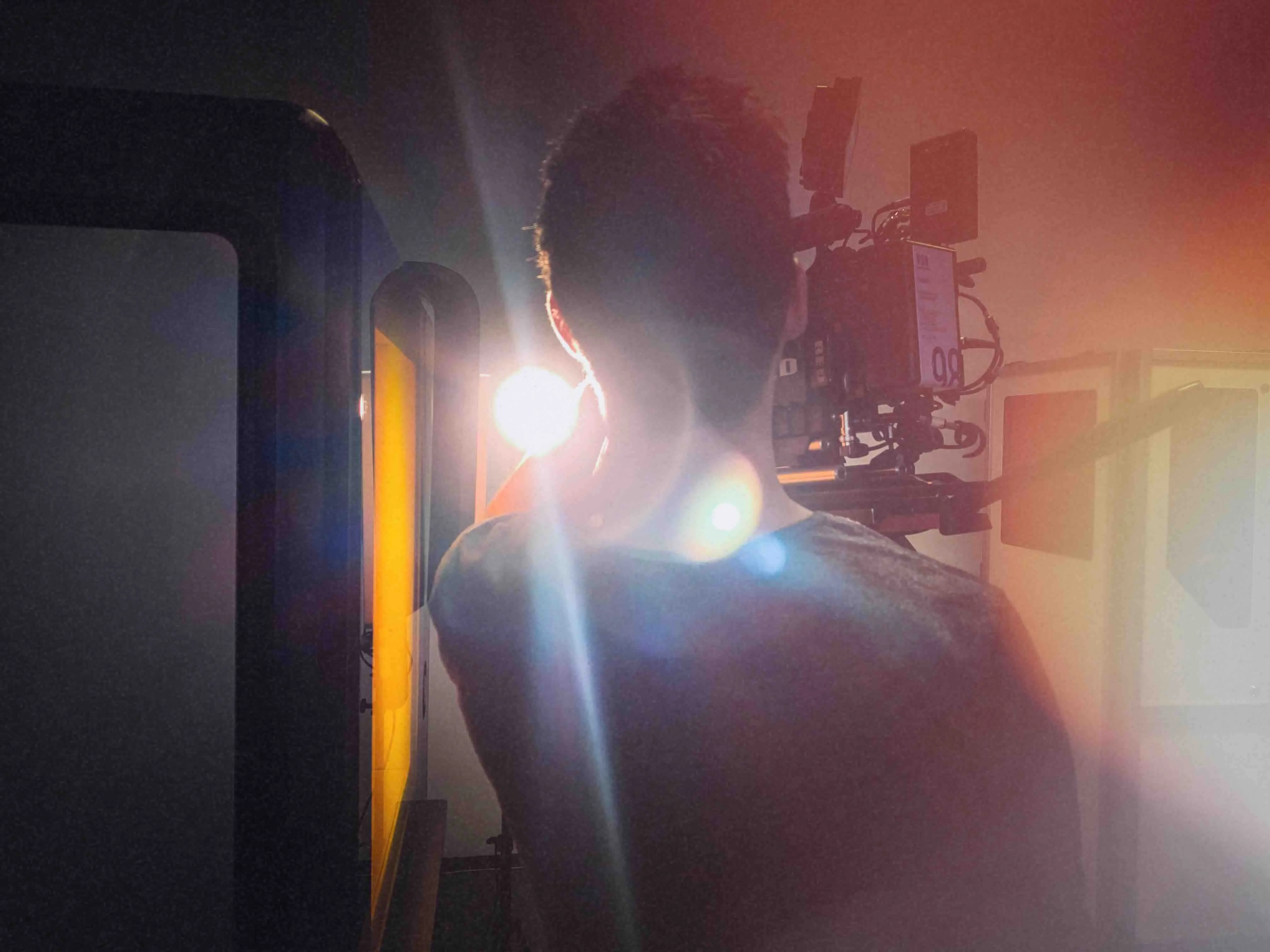 Person, von hinten gesehen, filmt mit einer Kamera, neben einer hellen Lichtquelle in einem dunklen Raum.