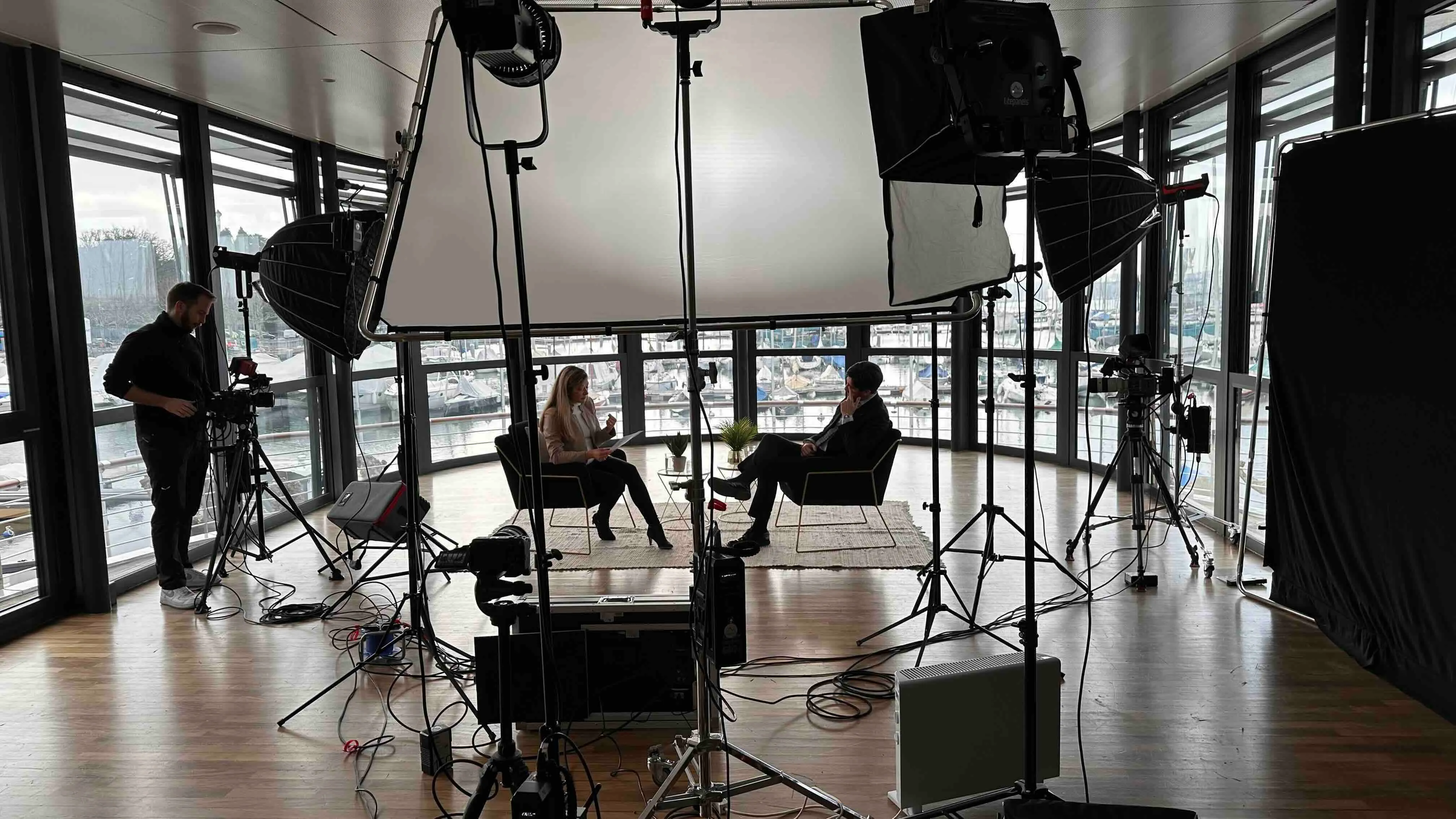 Zwei Personen führen ein Interview in einem modernen Raum mit großen Fenstern und Hafenblick, umgeben von Beleuchtungs- und Filmgeräten.