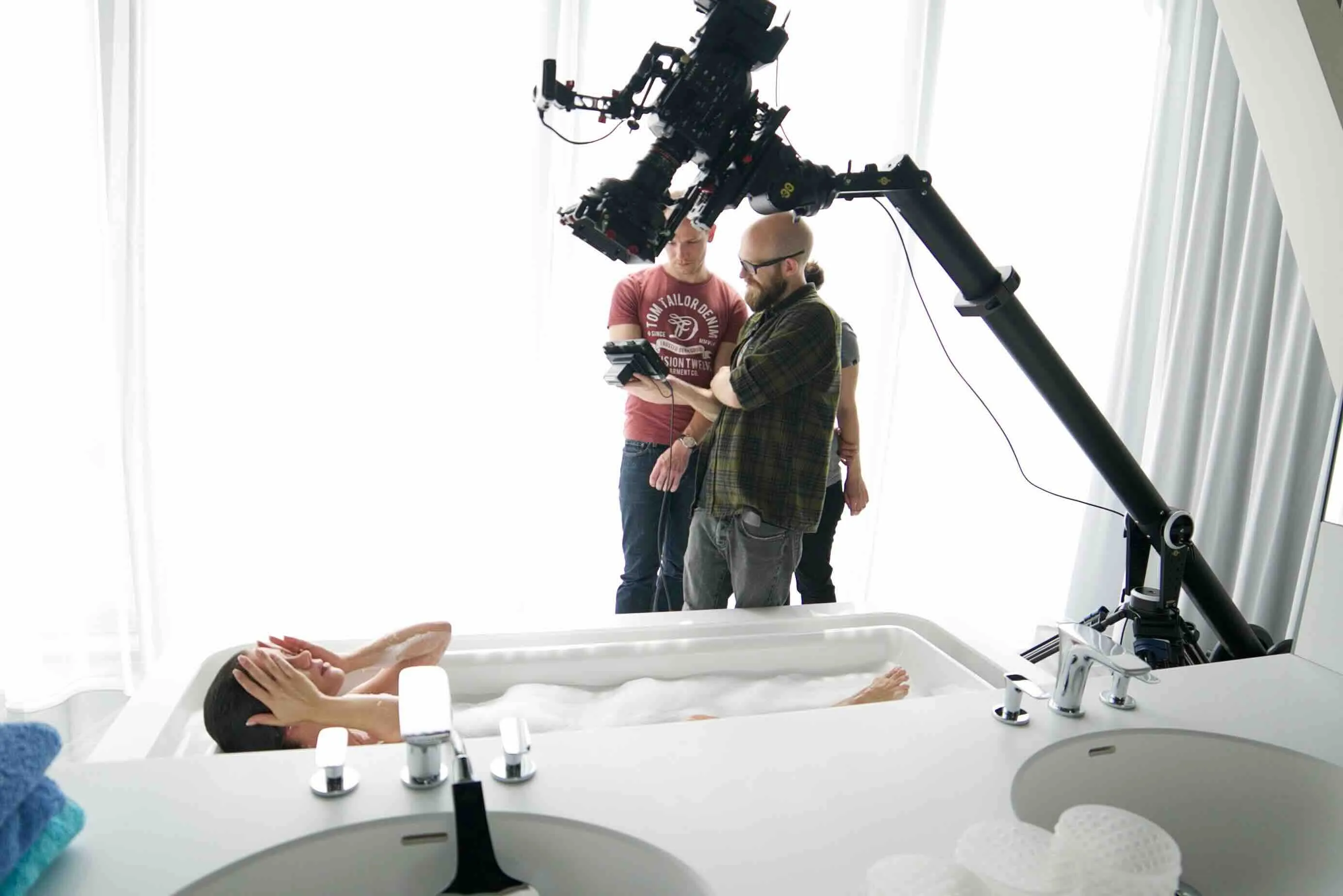 Eine Frau liegt entspannt in einer mit Schaum gefüllten Badewanne, während drei Männer hinter einer großen Kameraausrüstung stehen und Bildmaterial überprüfen.