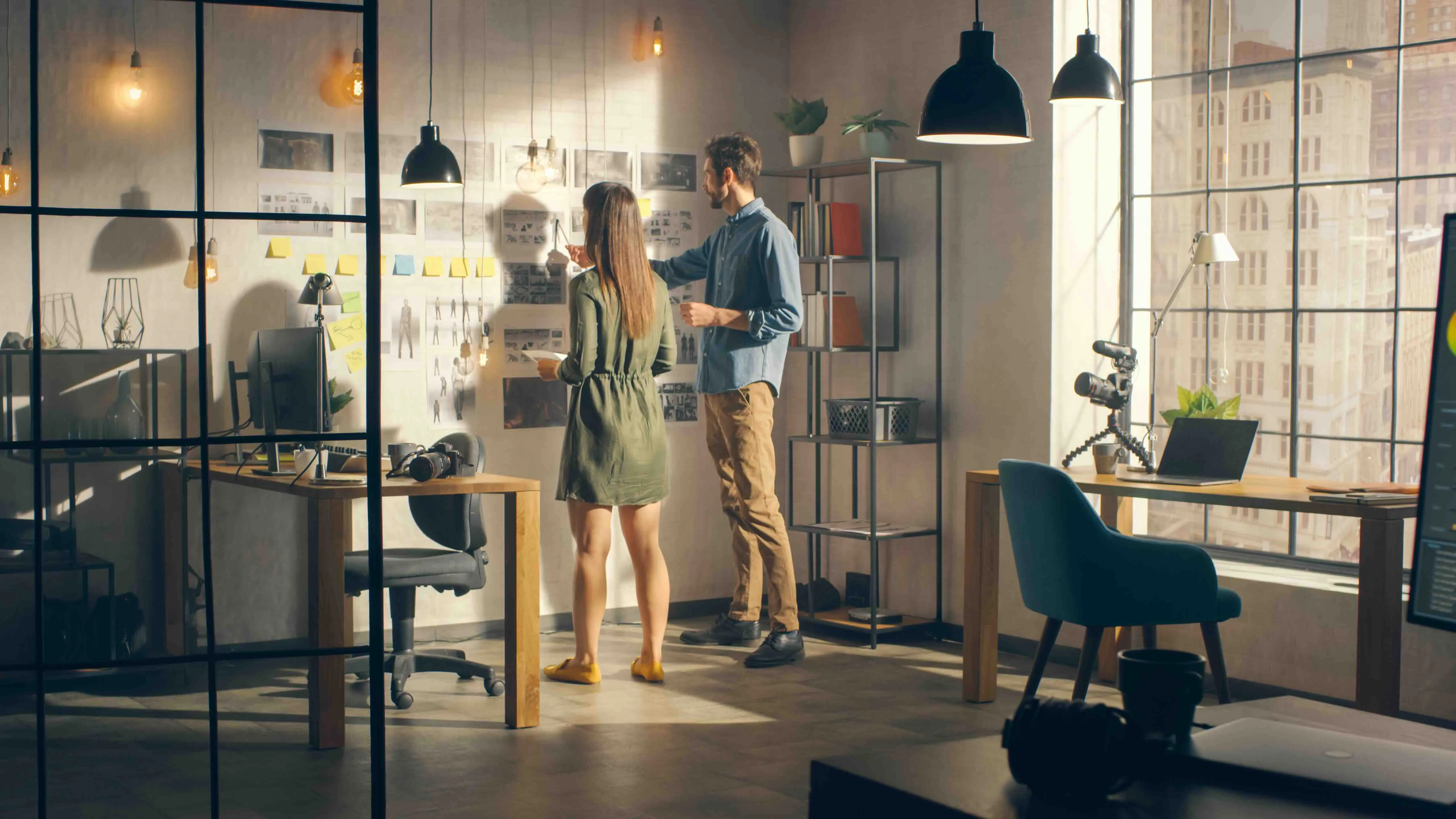 Zwei Personen besprechen kreative Skizzen und Notizen an einer Wand in einem modernen, hellen Büro mit großen Fenstern.