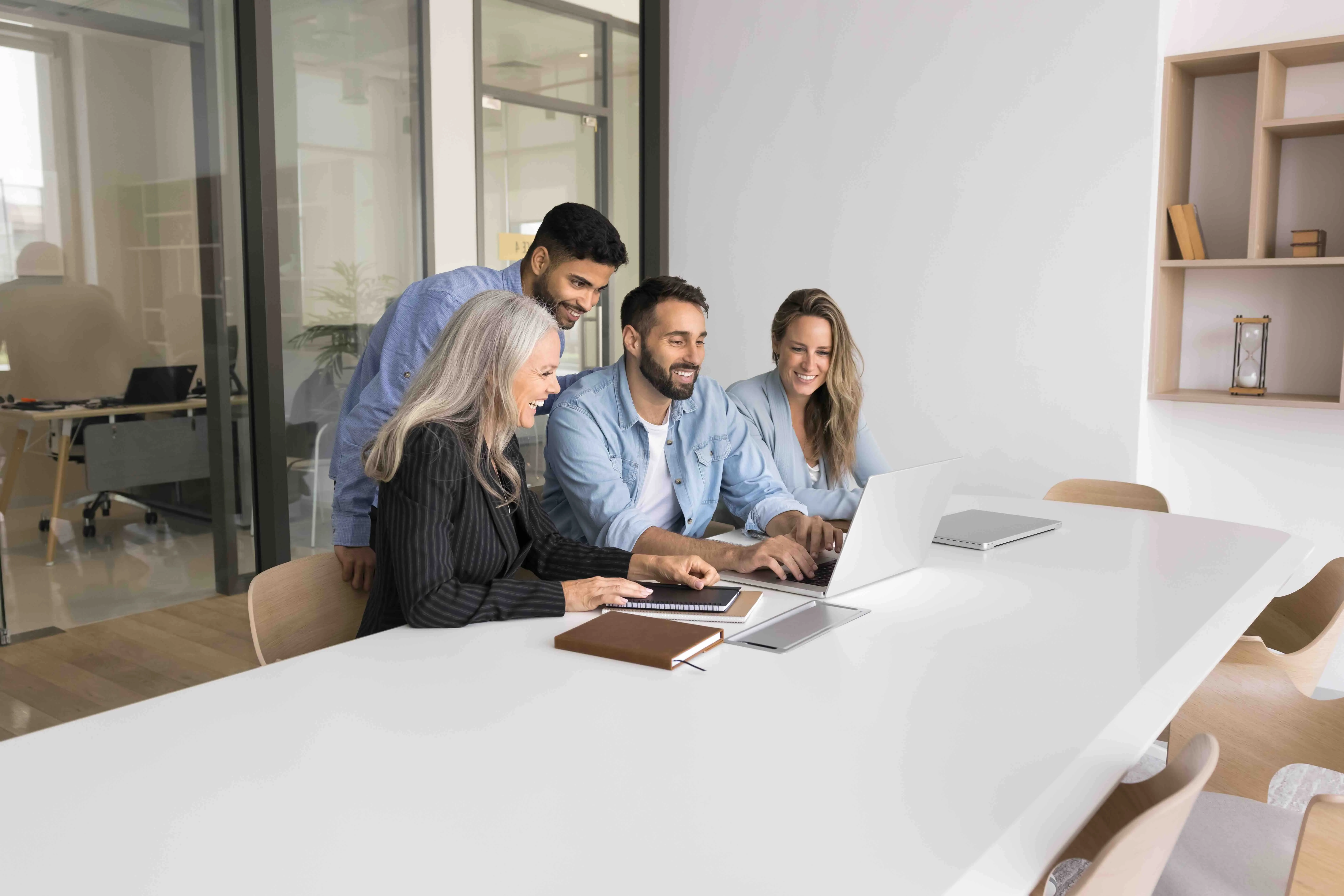 Vier Personen arbeiten gemeinsam lächelnd an einem Laptop in einem modernen Konferenzraum.