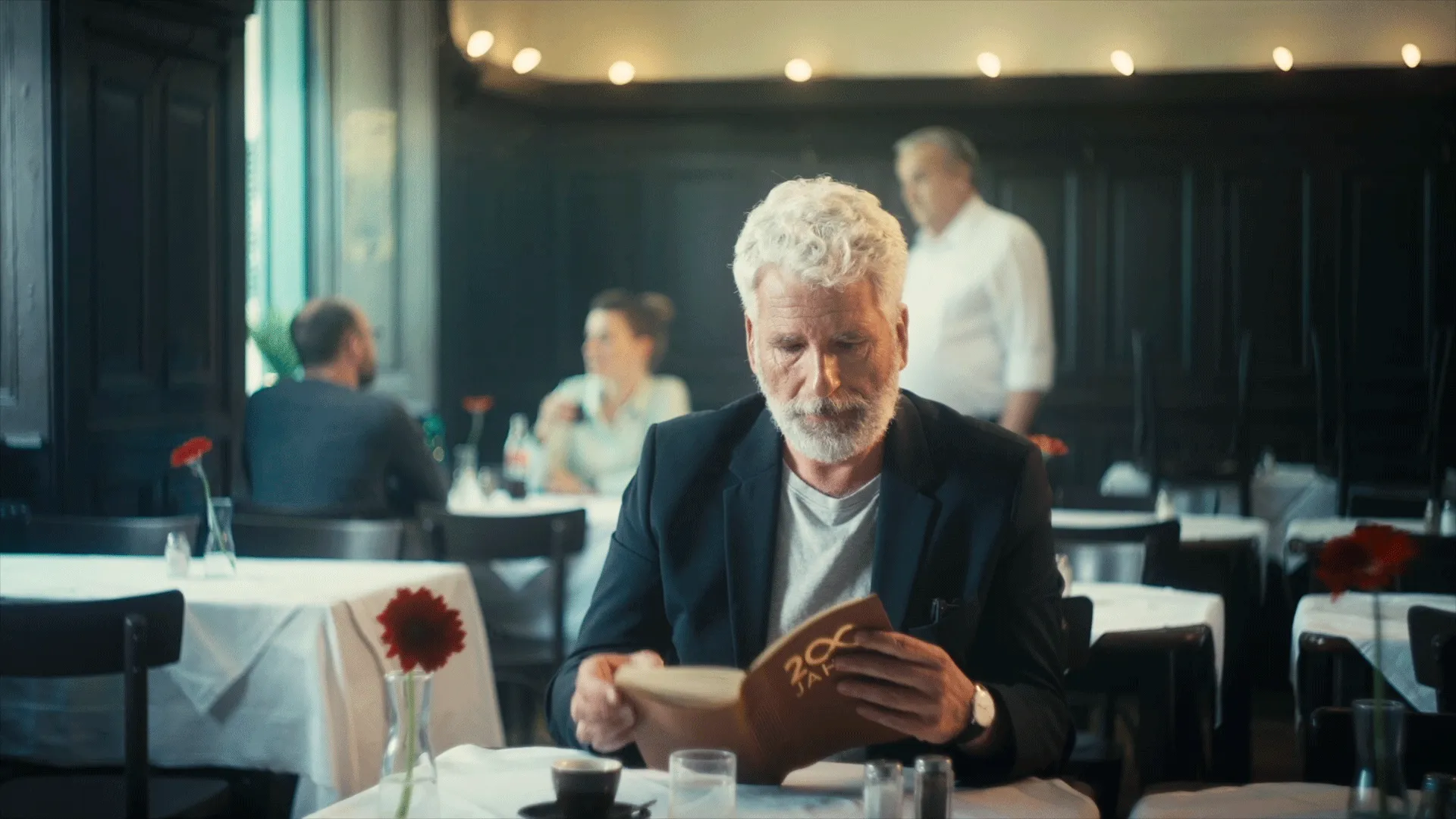 Älterer Mann mit weißem Bart und Locken liest ein Buch in einem Café mit weißen Tischdecken und roten Blumen.