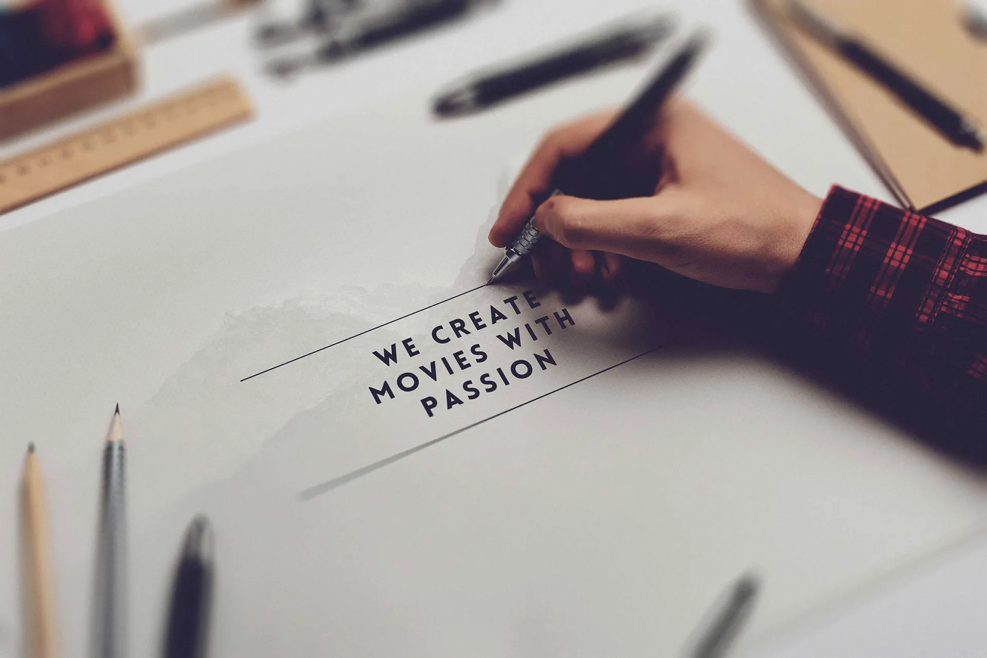 Hand schreibt mit Stift den Text „We create movies with passion“ auf weißem Papier, umgeben von Schreibutensilien.