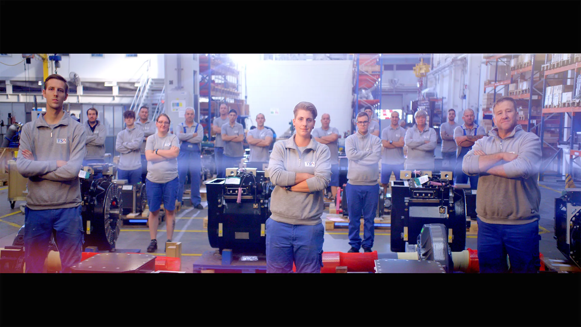 Team von Fabrikarbeitern steht in einer Produktionshalle mit verschränkten Armen vor Maschinen.