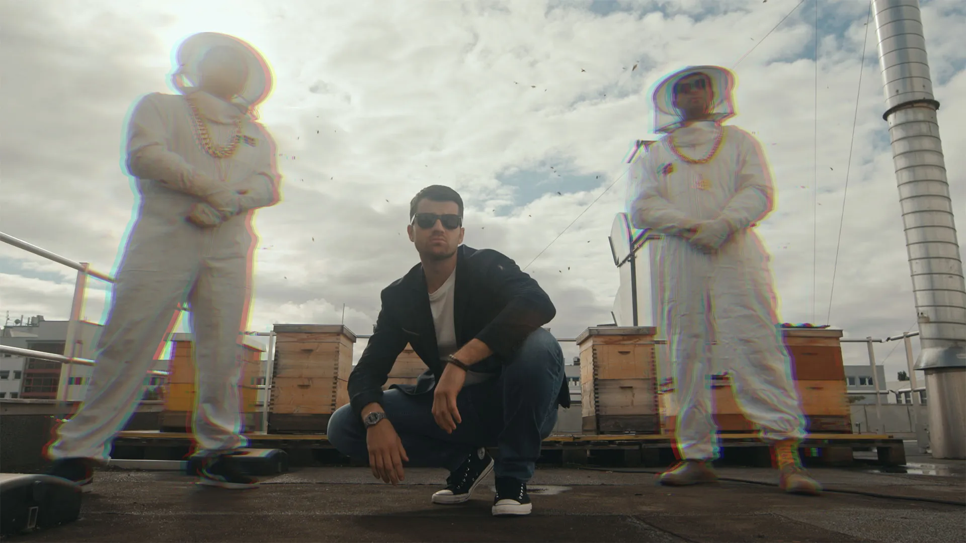 Junger Mann in Sonnenbrille hockt auf einem Dach zwischen zwei Personen in Bienenschutzausrüstung.