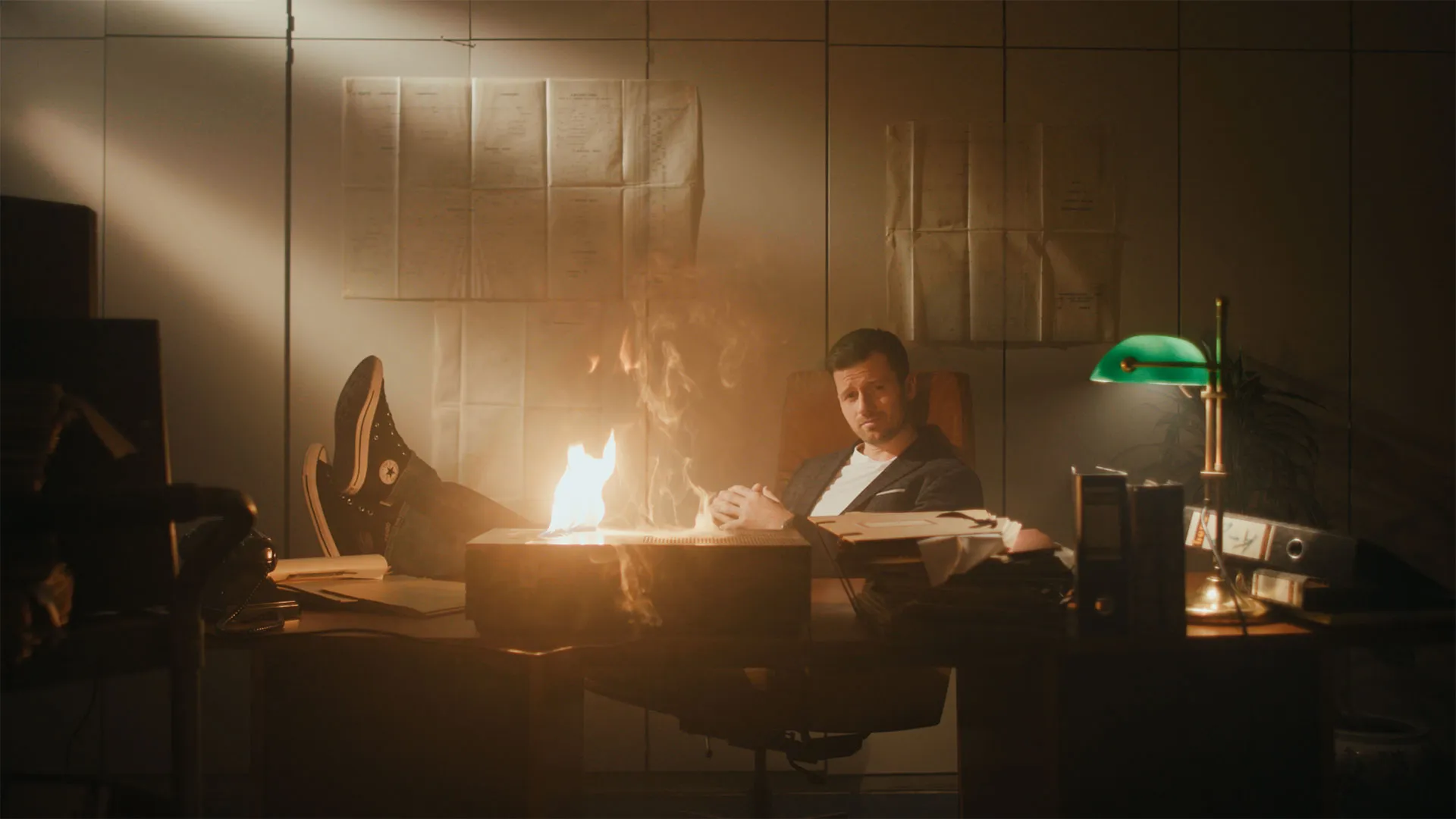 Mann lehnt zurück an seinem Schreibtisch mit brennendem Aktenordner, trägt schwarze Turnschuhe und schaut skeptisch.