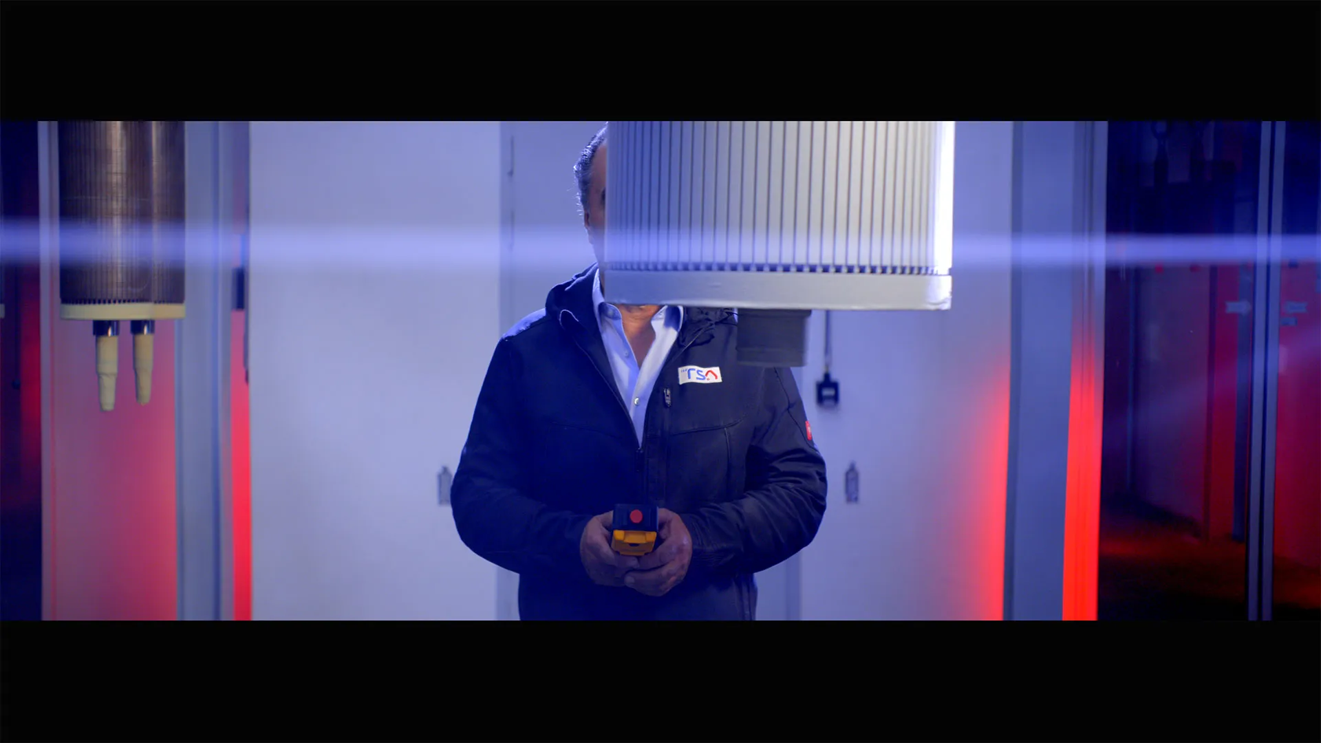 Mann in dunkler Jacke hält ein Gerät in der Hand, sein Gesicht wird von einer großen Maschinenkomponente verdeckt.