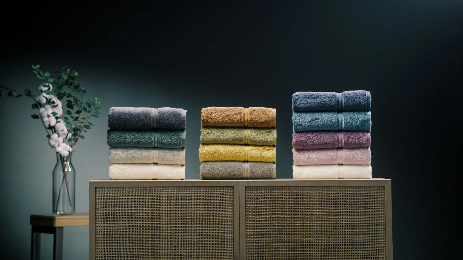 Drei Stapel farbiger Handtücher auf einem Holzschrank vor dunklem Hintergrund, daneben eine Vase mit Zweigen und weißen Baumwollblüten.