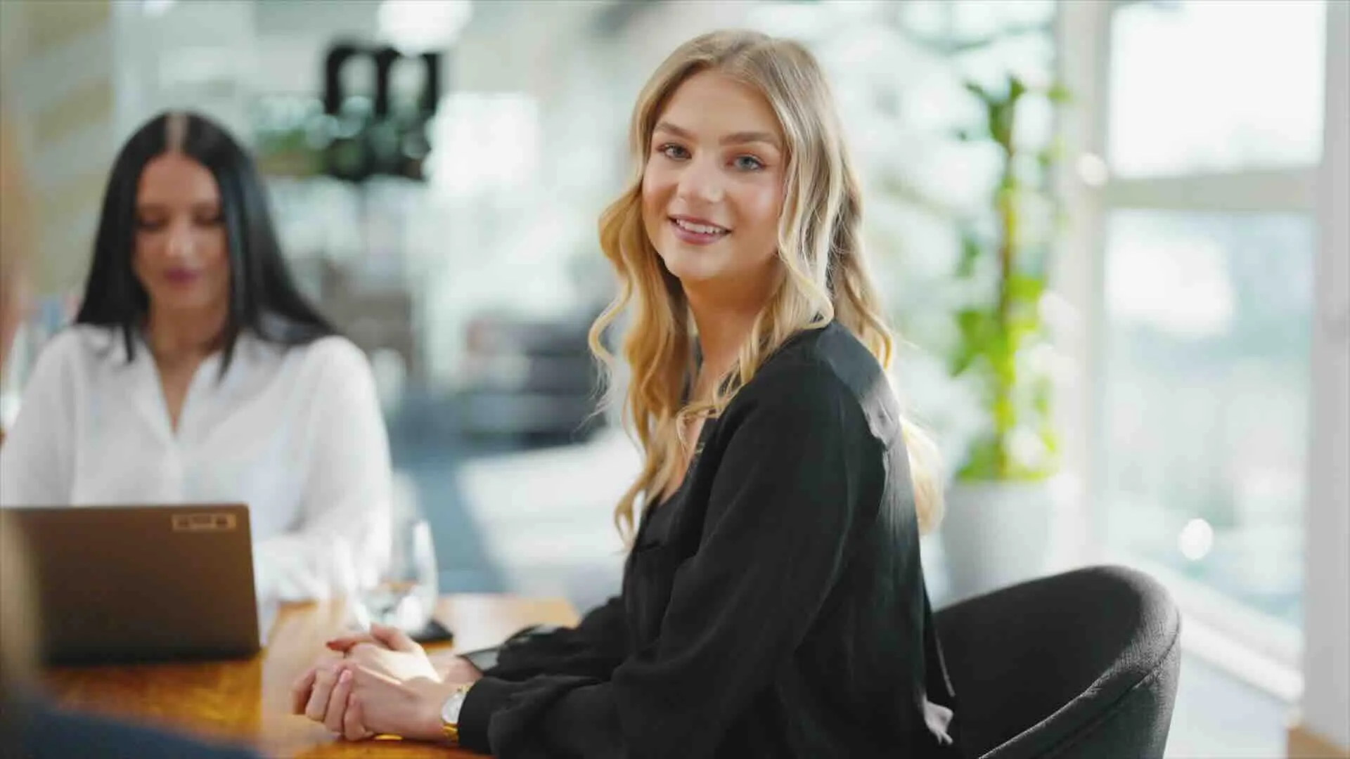 Junge blonde Frau in schwarzer Bluse sitzt an einem Tisch in einem modernen Büro und lächelt in die Kamera, im Hintergrund eine Frau mit langem schwarzem Haar, die an einem Laptop arbeitet.