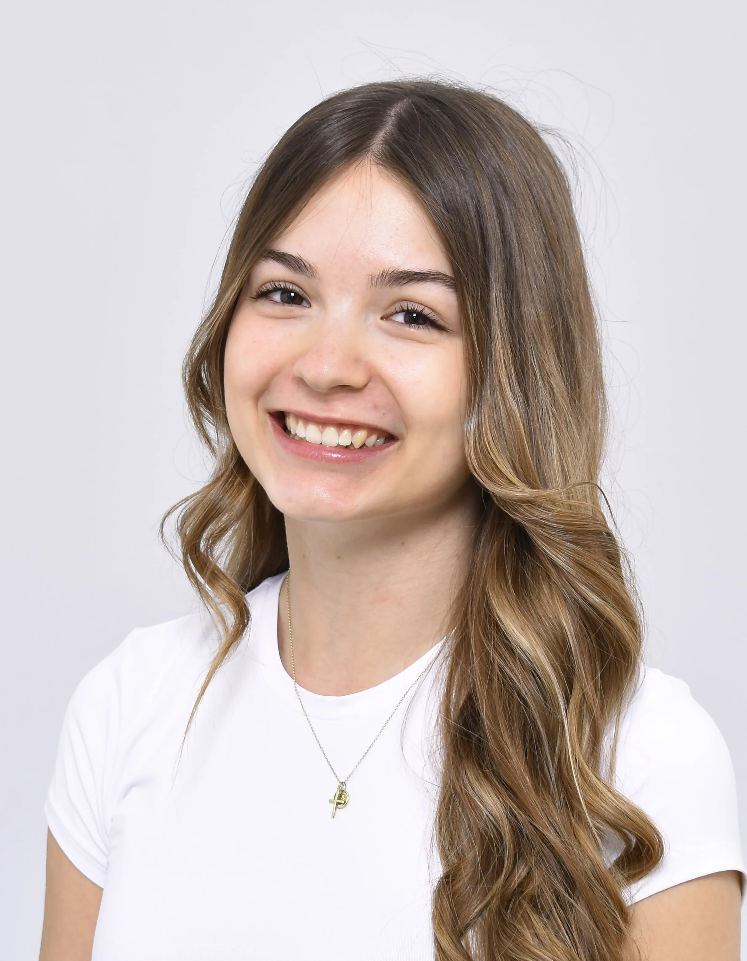 Junge Frau mit langen welligen braunen Haaren und weißem T-Shirt lächelt vor hellem Hintergrund.