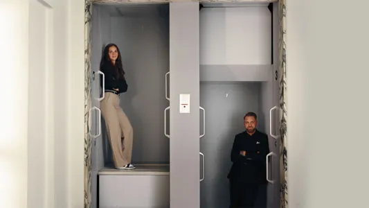 Eine Frau steht links im Aufzug mit verschränkten Armen, ein Mann steht rechts im Aufzug vor einer hellgrauen Wand, beide schauen in die Kamera.