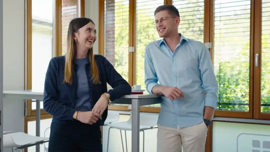 Zwei Personen stehen in einem hellen Raum an einem hohen Tisch und unterhalten sich lächelnd.