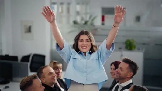 Junge Frau hebt freudig die Arme, umgeben von lachenden Kollegen in einem Büroumfeld.