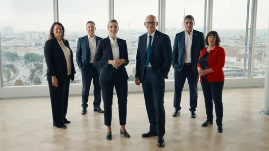 Sechs Geschäftsleute in formeller Kleidung stehen in einem modernen Büro mit großen Fenstern und Stadtblick.