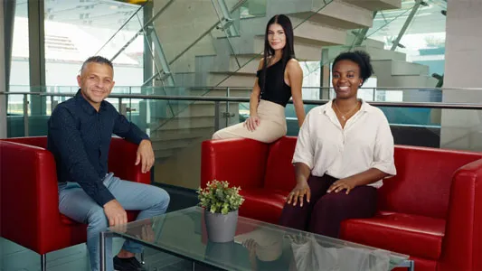 Drei lächelnde Personen, zwei Frauen und ein Mann, sitzen auf roten Sofas in einem modernen Innenraum mit Glastisch und Treppe im Hintergrund.