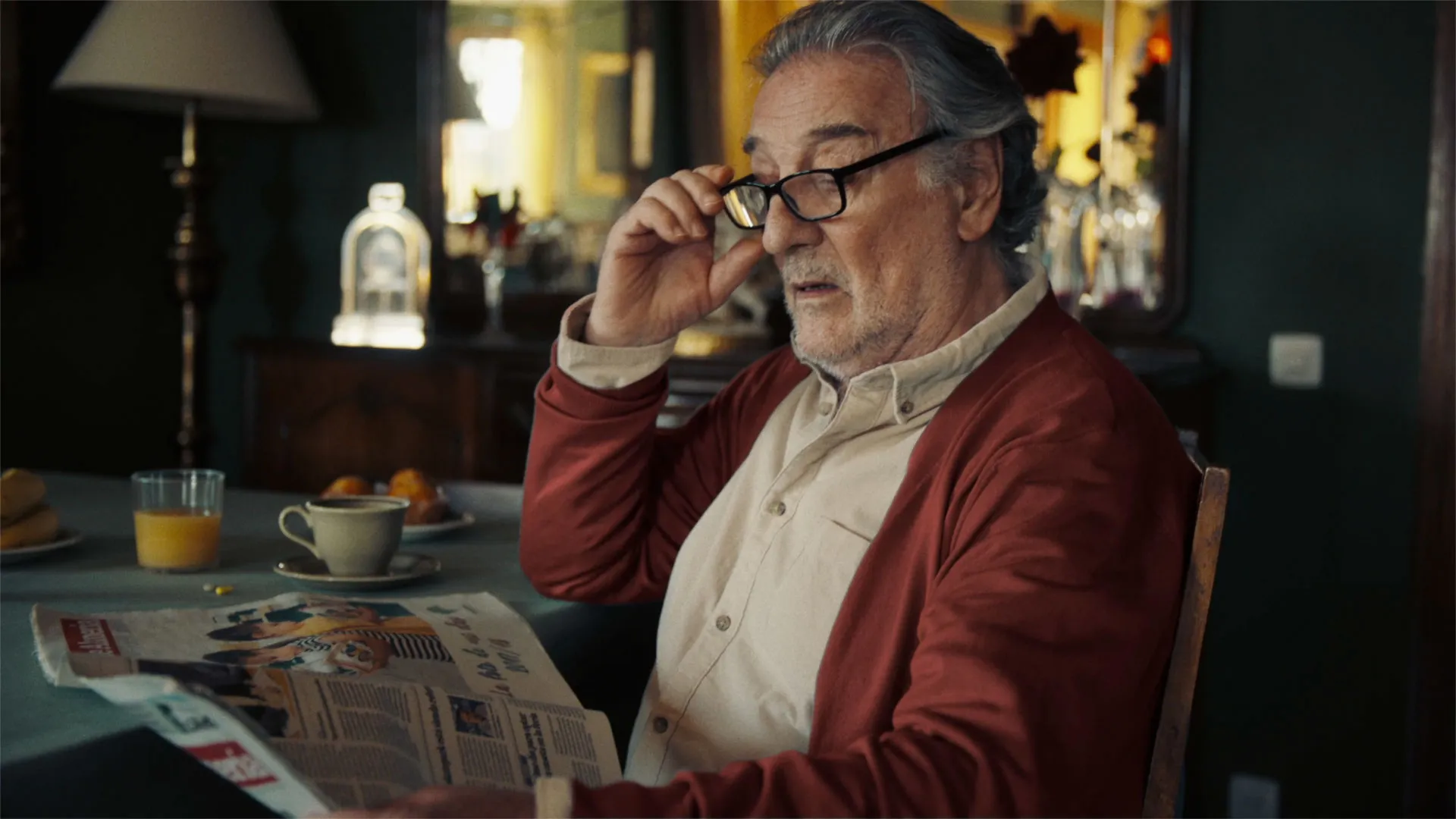 Elderly man with glasses sitting at a table and reading a newspaper.