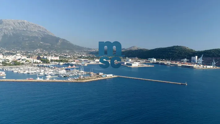 Hafen mit vielen Booten vor einer Küstenstadt, umgeben von Bergen und klarem blauem Himmel.