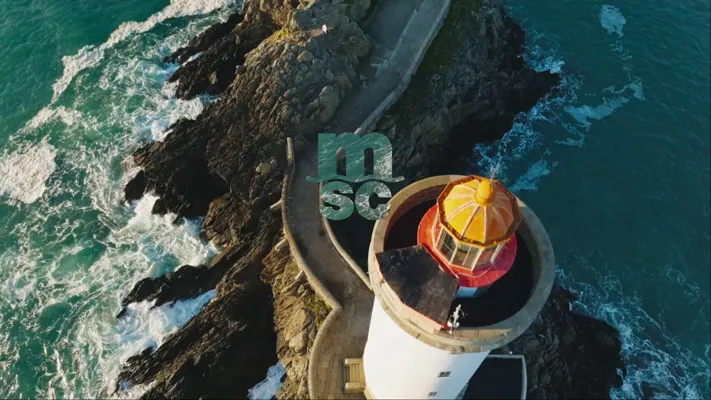 Drohnenaufnahme eines weißen Leuchtturms mit gelber Kuppel auf felsiger Küste mit blauem Meer.