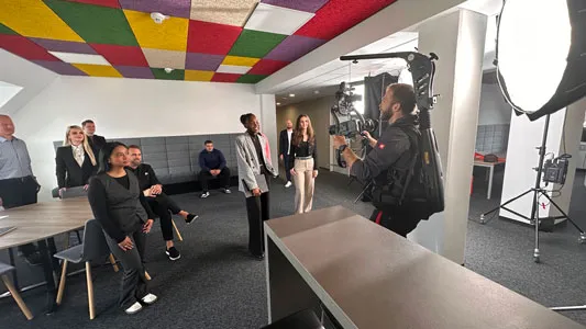 Ein Kameramann filmt in einem Büro ein Team von Personen, von denen einige sitzen und andere stehen, vor einer Wand mit farbigen Akustikplatten an der Decke.