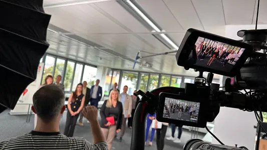 Eine Person vor einer Kamera filmt eine Gruppe von Menschen in einem hellen, modernen Büro.