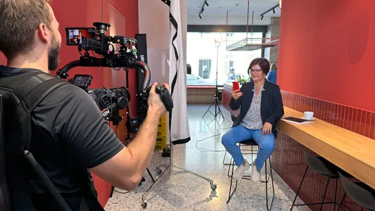 Eine Frau sitzt auf einem Hocker vor roter Wand in einem Café und hält ein rotes Smartphone, während ein Kameramann sie mit professioneller Kamera filmt.