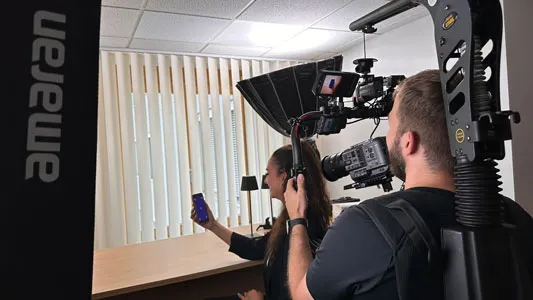 Kameramann filmt Frau mit Smartphone vor Jalousien und Lampe.