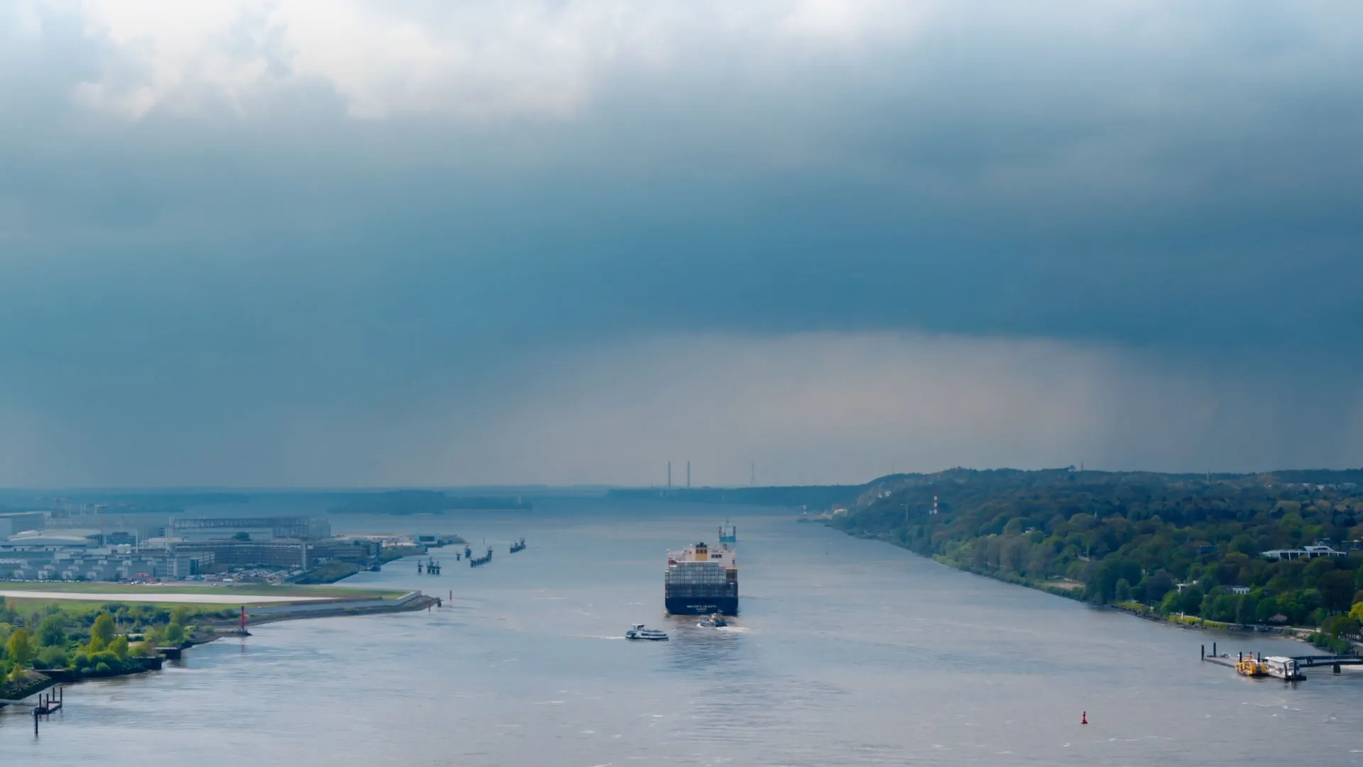 Frachtschiff fährt auf einem breiten Fluss unter bewölktem Himmel, mit bewaldetem Ufer auf der rechten Seite und Industrieanlagen links.