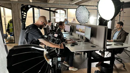 Film crew producing an image film in a modern office with camera, light setup and actor in front of the computer.