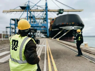 Video production at a port: film crew in safety vests filming scenes in front of a large container ship.