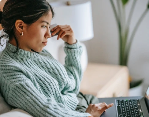 A woman sat on her sofa with her laptop, holding her bank card looking concerned about paying her mortgage. 