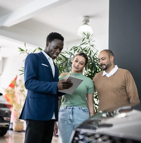 A car salesman using a digital tablet while talking about Tandem Motor Finance to a young couple looking to purchase a new lower emission car on finance.