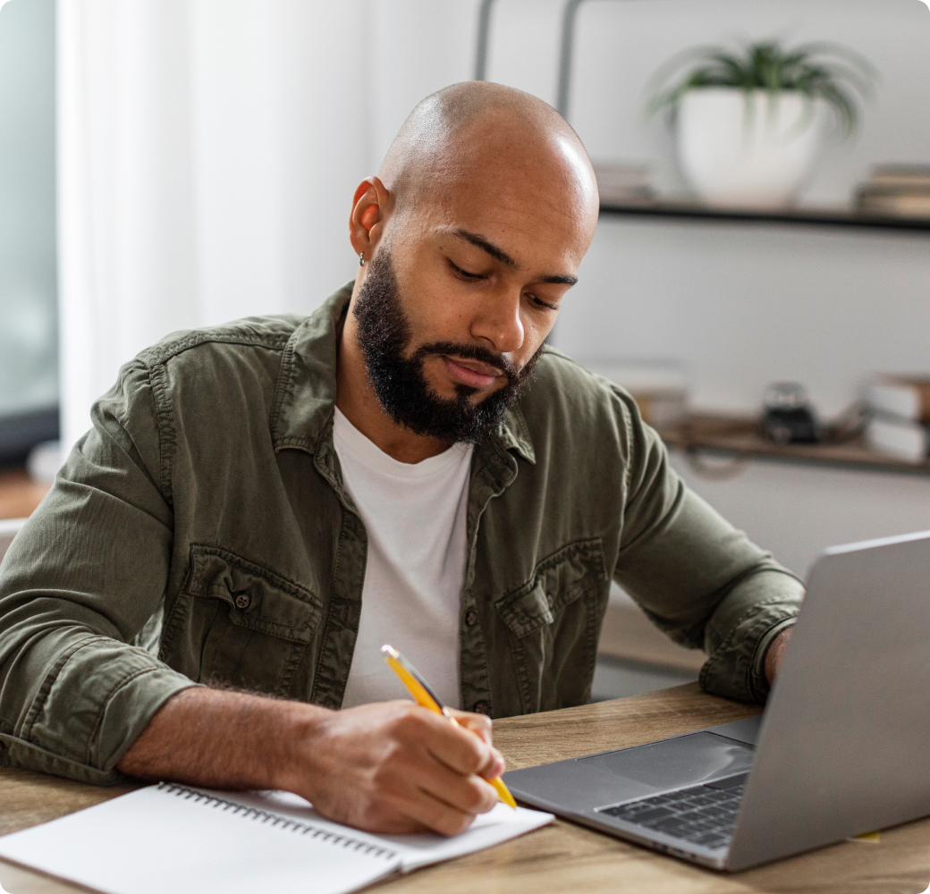 A young man, visibly affected by financial hardship, sits at his dining table, poised with pen and paper, preparing to reach out to Tandem regarding his current financial challenges impacting his loan repayments.