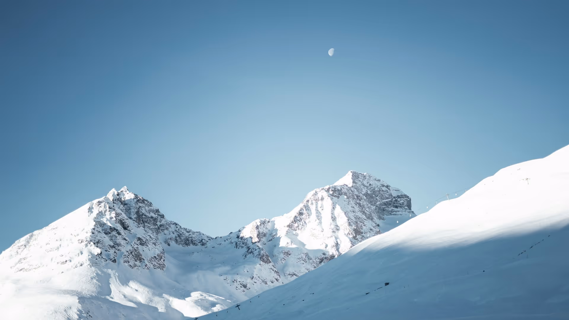 Schneebedeckte Berggipfel unter blauem Himmel mit sichtbarem Halbmond.