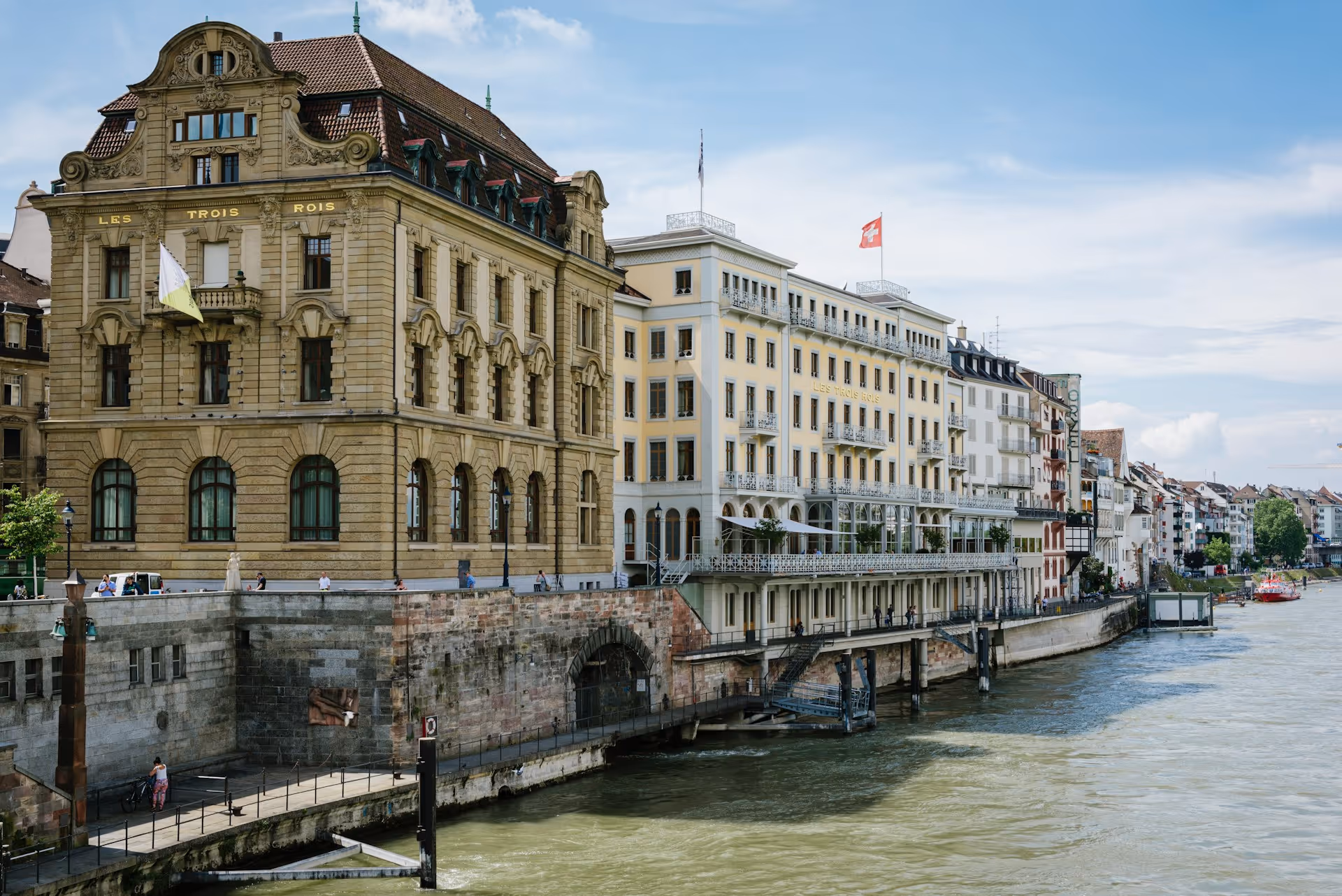 Blick auf historische Gebäude entlang eines Flussufers mit Schweizer Flagge und Menschen, die am Ufer entlang spazieren.