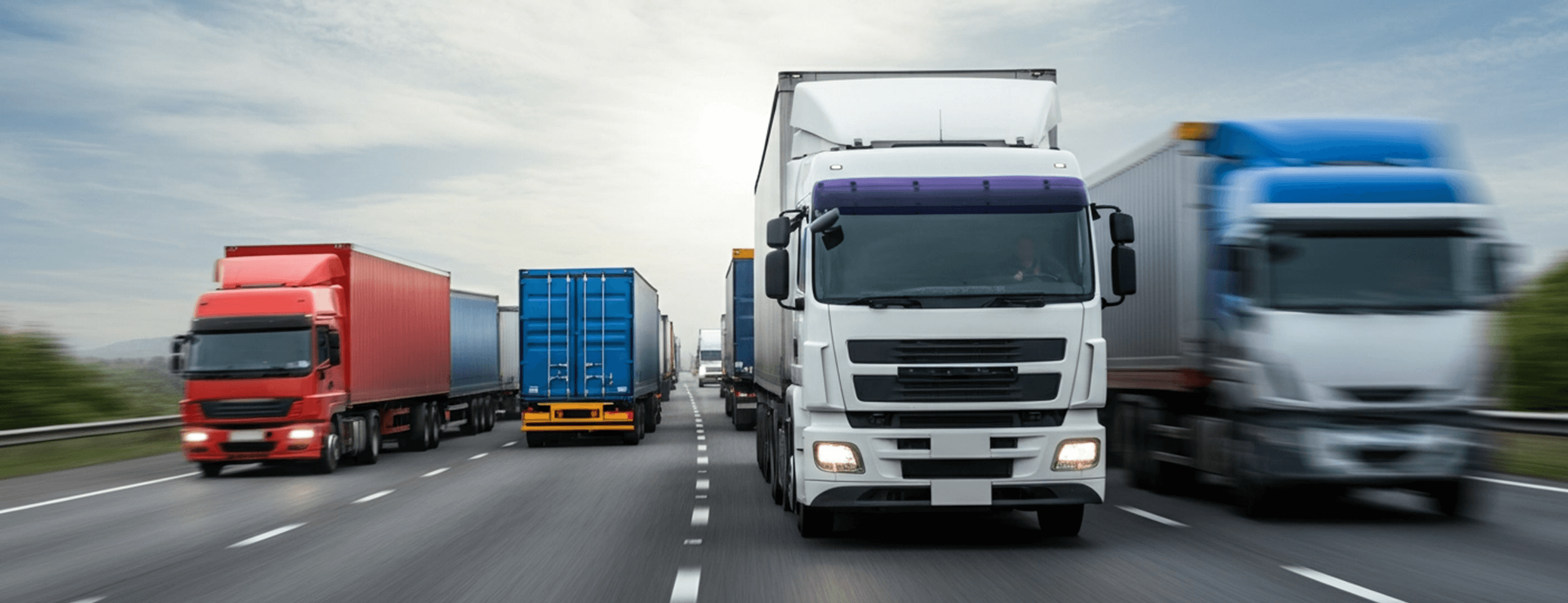 Several cargo trucks, including a prominent white one, driving on a highway under a cloudy sky.