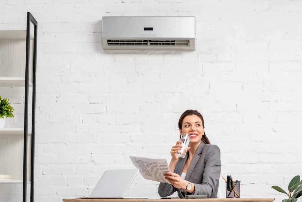A professional woman sits at a desk, smiling while holding a glass of water, with an indoor wall-mounted AC unit visible above her.