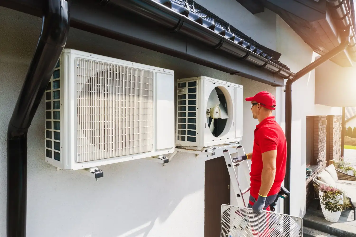  A technician wearing a red shirt, red cap, and safety glasses is working on two white outdoor heat pump condenser units mounted on the wall of a house under a dark gutter. He is standing on a ladder, holding a fan guard panel for the unit on the right, which has its fan exposed. Sunlight shines from the right.