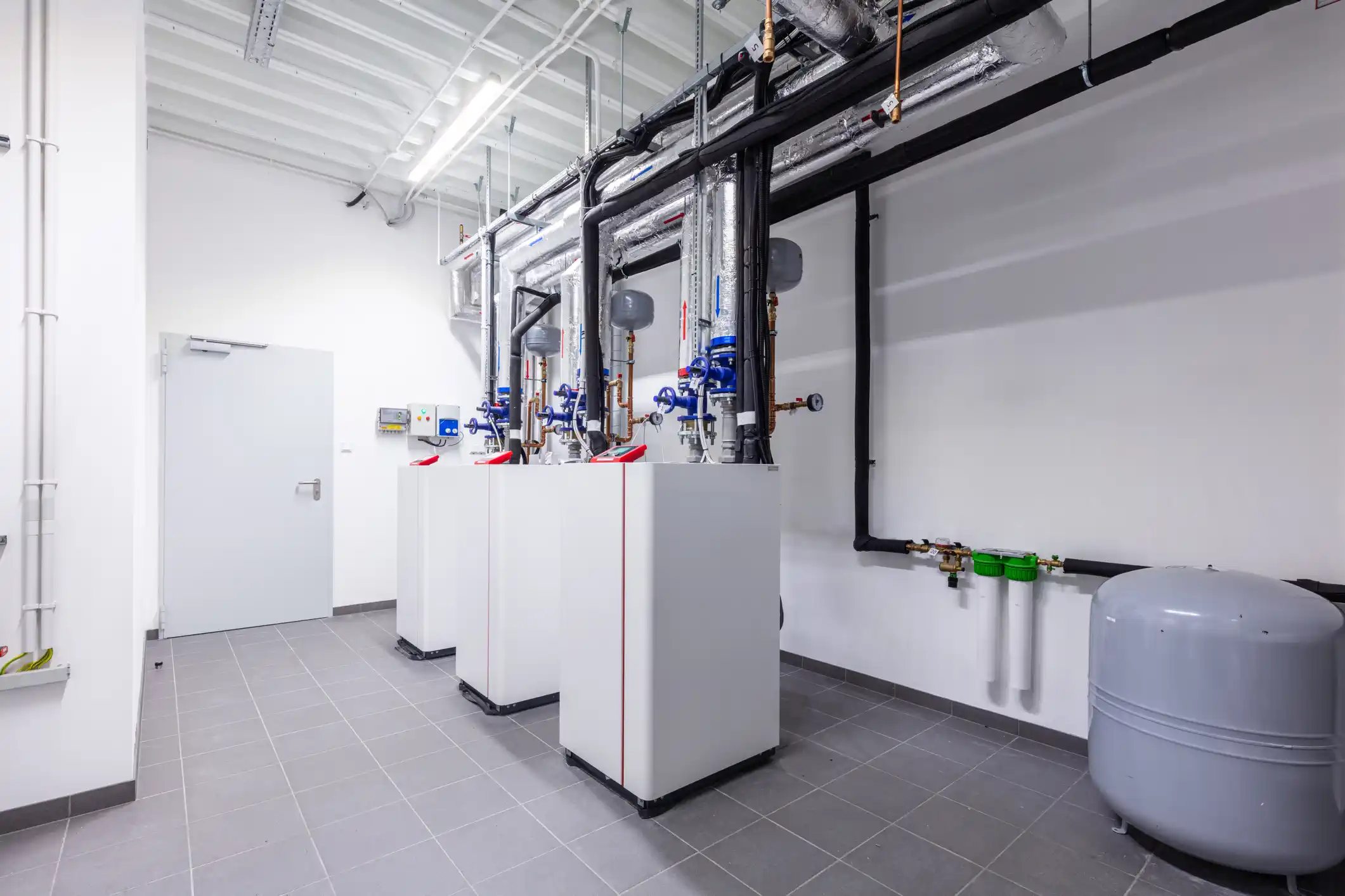  Three white, modern commercial boiler or water heater units with red accents are installed in a clean utility room with white walls and a gray tiled floor. The units are connected to a complex network of black insulated pipes and manifolds mounted high on the wall, including an expansion tank and water filters on the right.