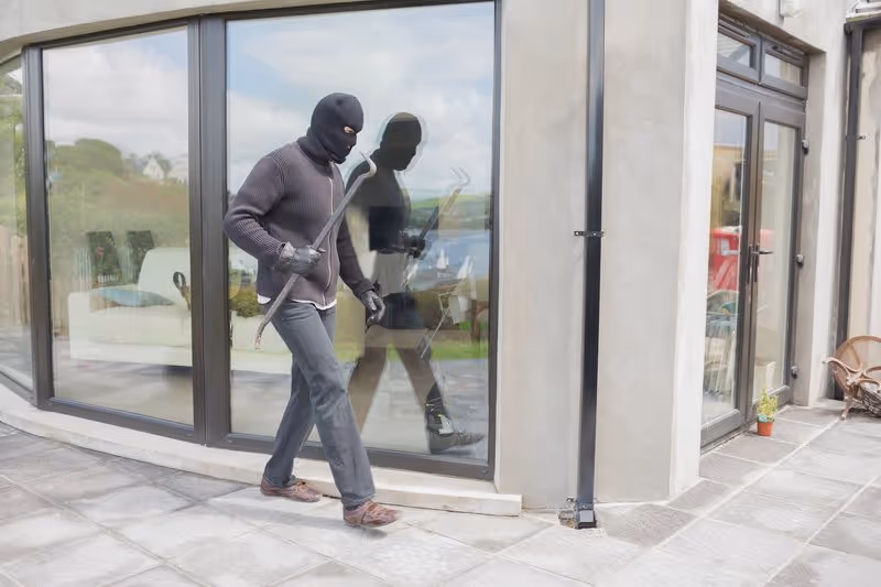 Cambrioleur masqué avec pied-de-biche tentant d'entrer dans une maison devant une baie vitrée.